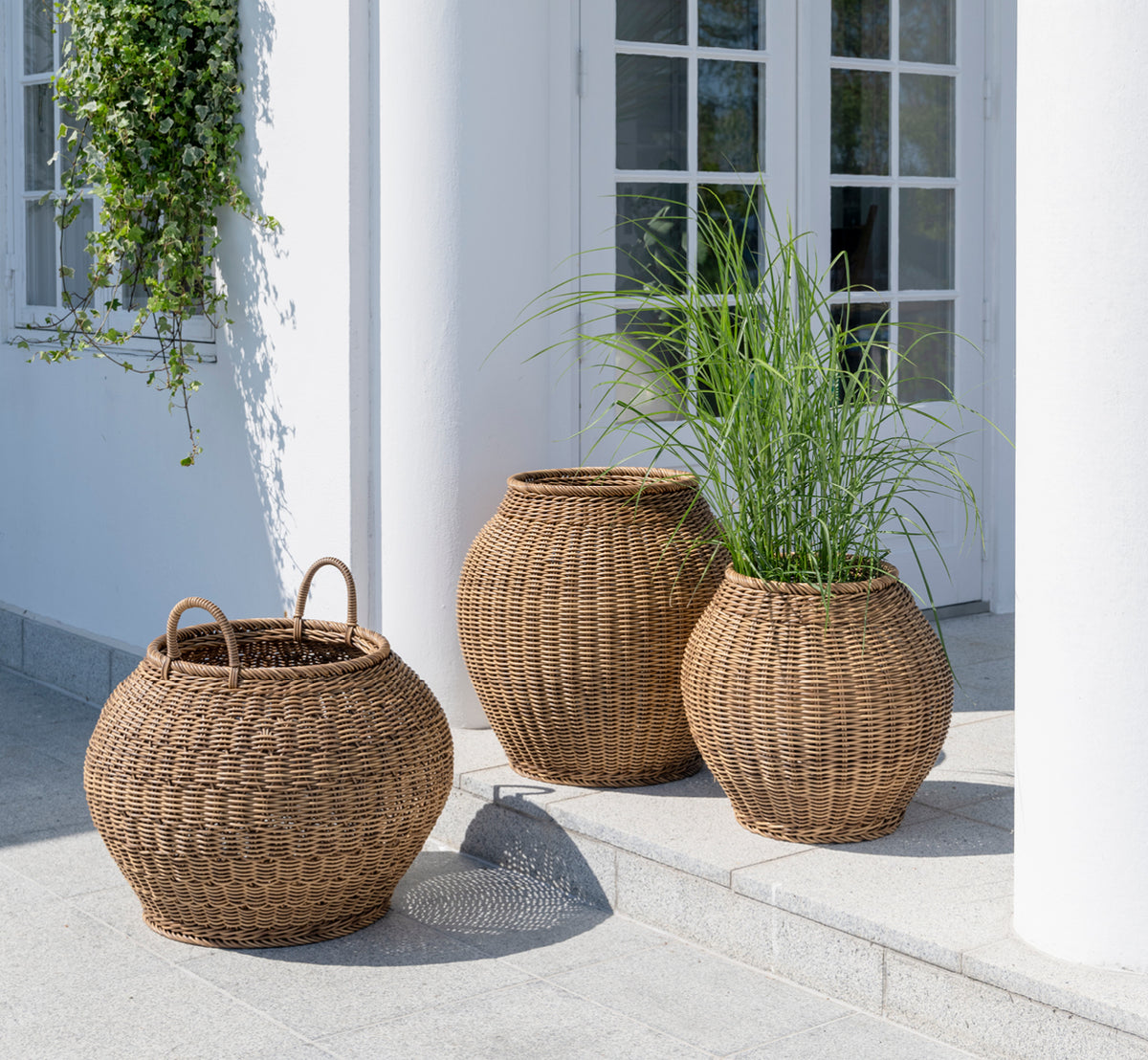 Dette er et livsstilsbillede af Ellis Kurv i chestnut core weave ArtFibre Aluminium, udendørs på en solbeskint terrasse med hvid mur og flisegulv.