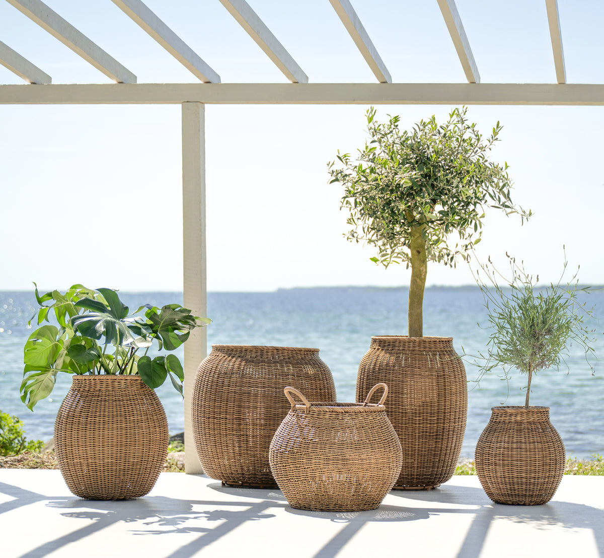 Dette er et livsstilsbillede af Ellis Kurv i chestnut core weave ArtFibre Aluminium, udendørs på en solbeskint terrasse med hvid mur og flisegulv.