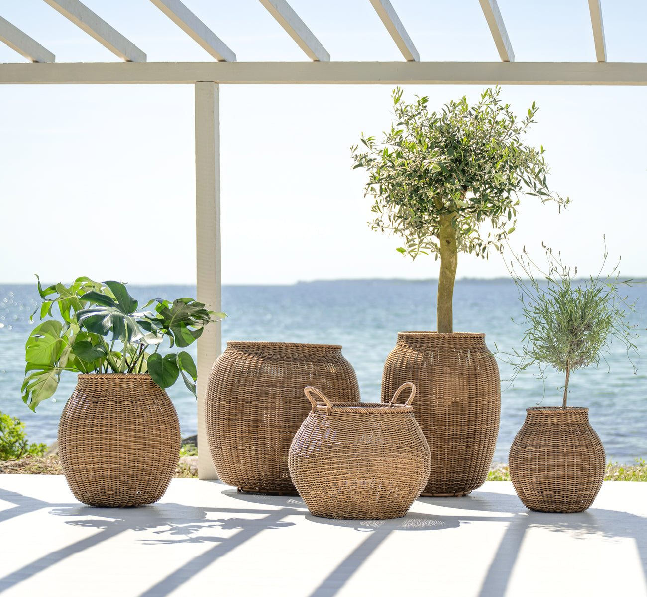 Dette er et livsstilsbillede af Ellis Kurv i chestnut core weave ArtFibre Aluminium, udendørs på en solbeskint terrasse med hvid mur og flisegulv.