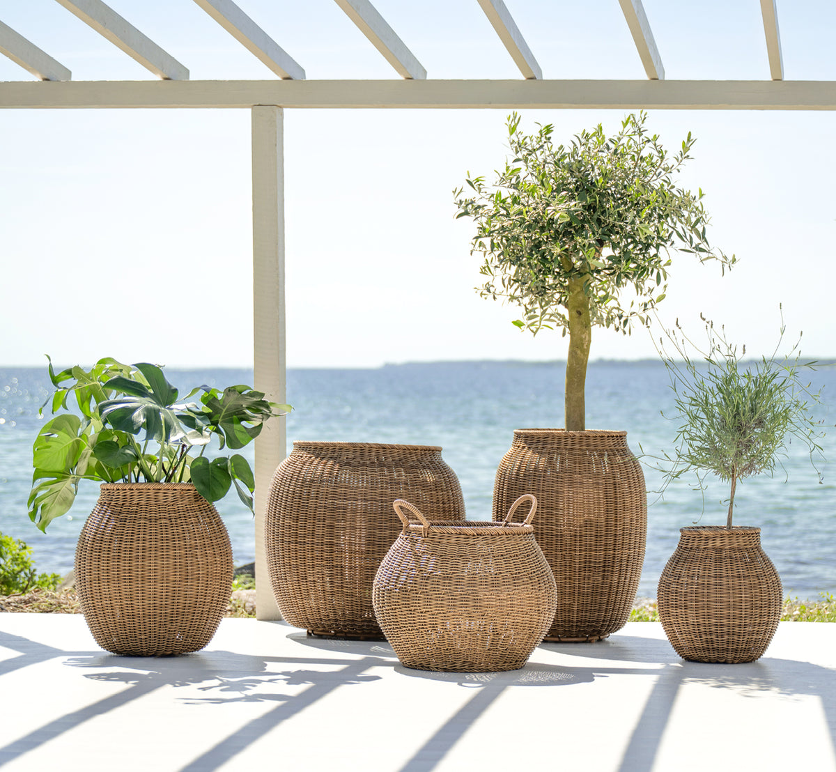 Dette er et livsstilsbillede af Ellis Kurv i chestnut core weave ArtFibre Aluminium, udendørs på en solbeskint terrasse med hvid mur og flisegulv.