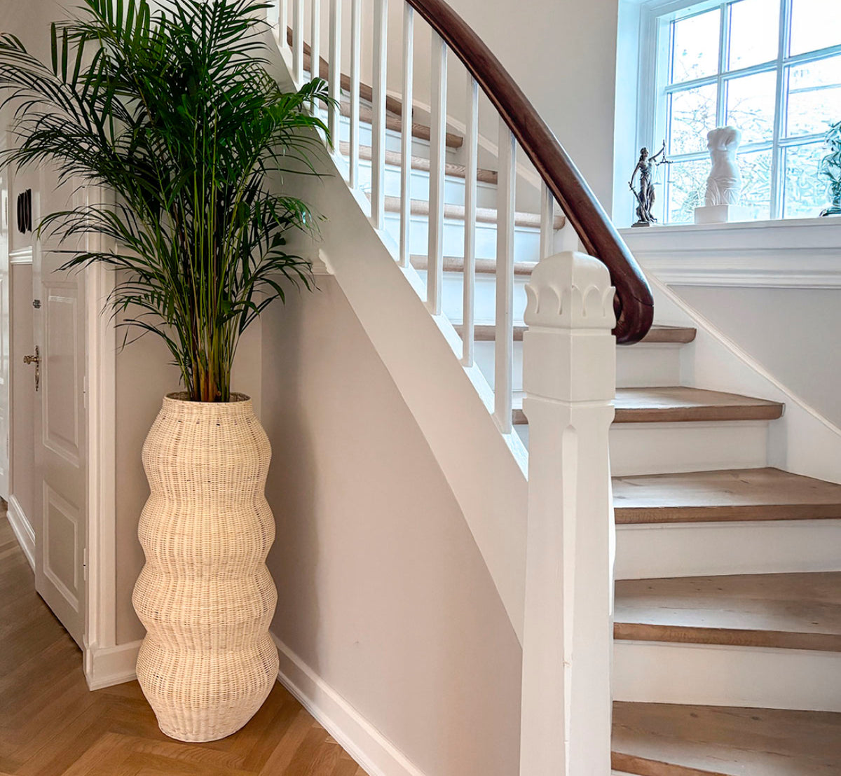 Dette er et livsstilsbillede af Unda Basket H102 cm i natural core weave iron frame. Brugt som en høj plantekumme ved siden af en buet trappe i en lys hall.