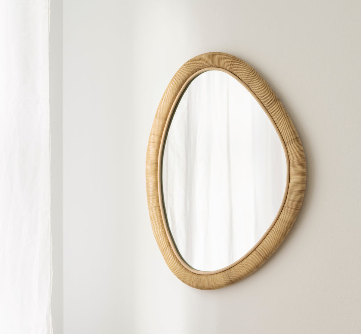 Dette er et livsstilsbillede af Malou Spejl i natural rattan. Organisk oval form monteret på en blød hvid væg med gardin.
