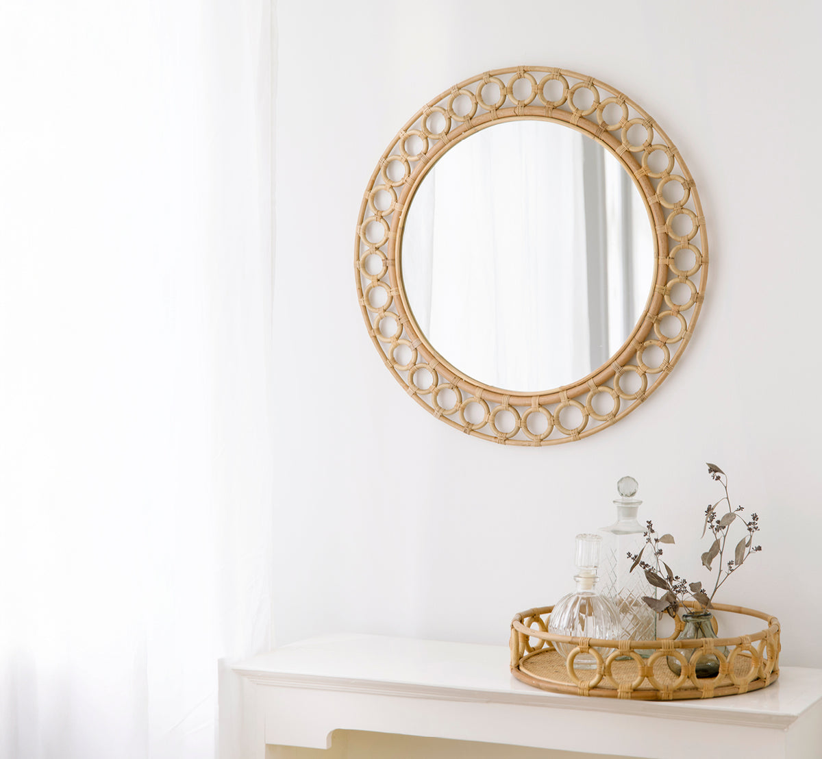 Dette er et livsstilsbillede af Mira Spejl i natural rattan. Monteret over en hvid konsol stylet med karafler og tørrede grene.