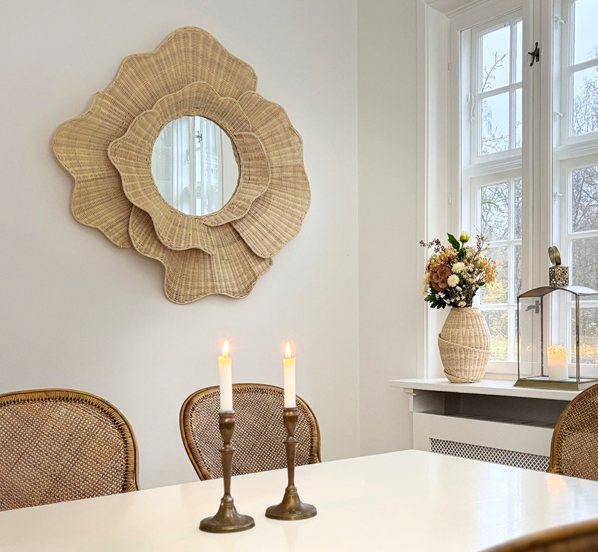 Dette er et livsstilsbillede af Floral Spejl i natural core weave iron frame, hængende i en lys spisestue med lysestager og naturligt vindueslys.