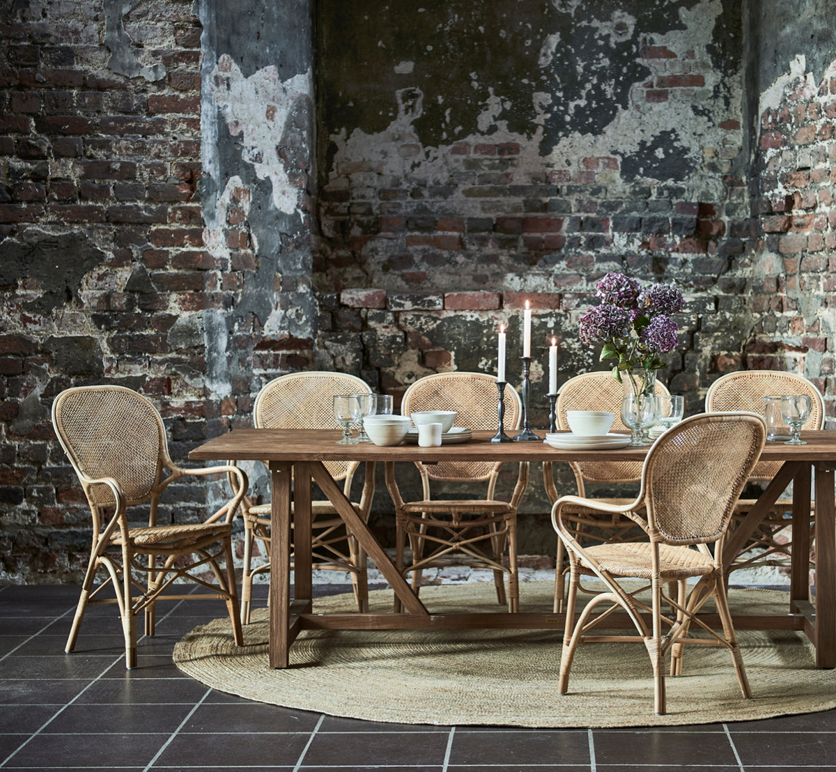 Dette er et livsstilsbillede af Jute Carpet Ø280 i natural jute. Under et spisebord med rattanstole i et rustikt rum med synlige murstensvægge.
