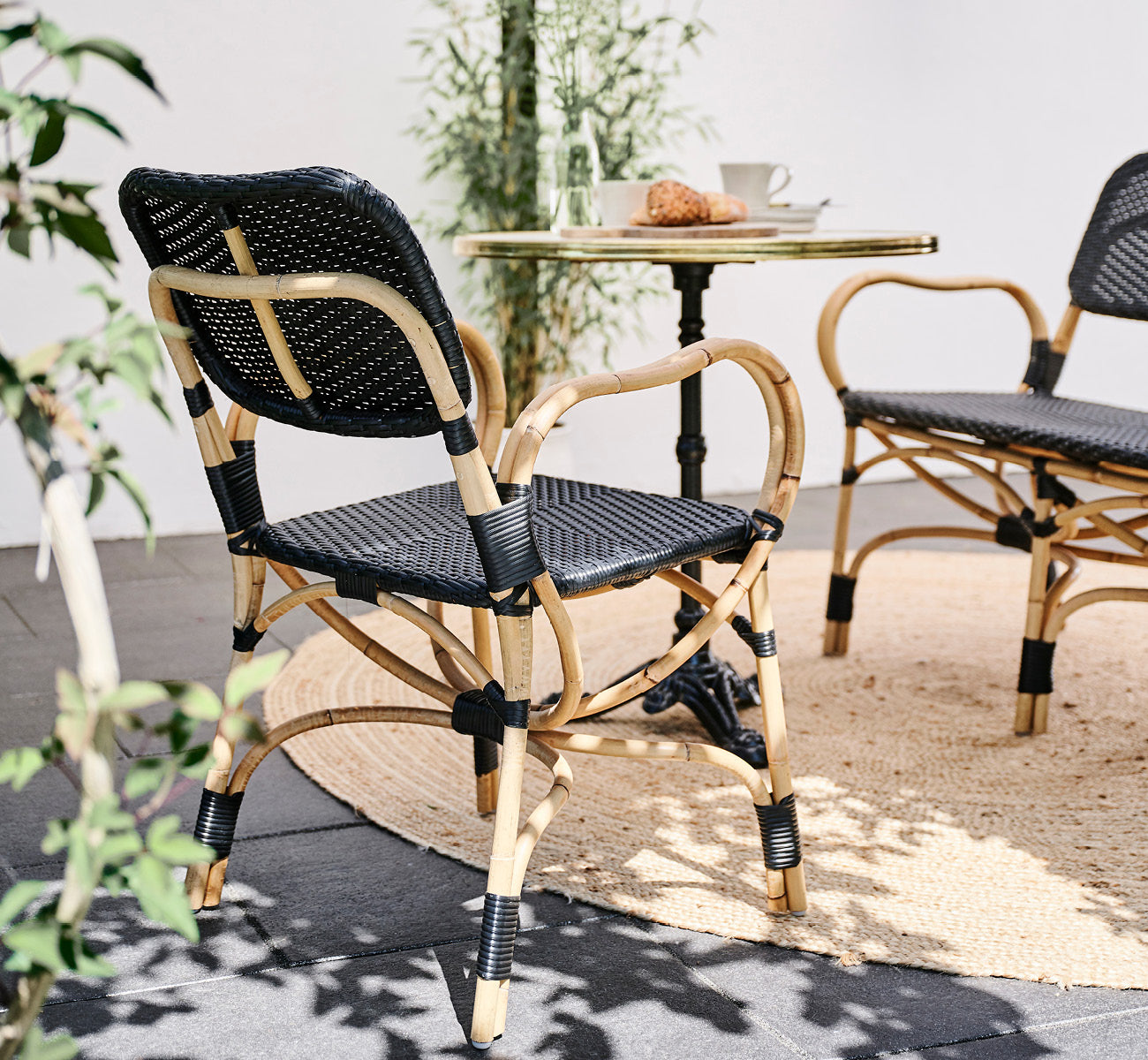 Dette er et livsstilsbillede af Bistro Dining Chair i skin on Rattan frame, i et udendørs café-miljø med et bistrobord på mørke fliser.