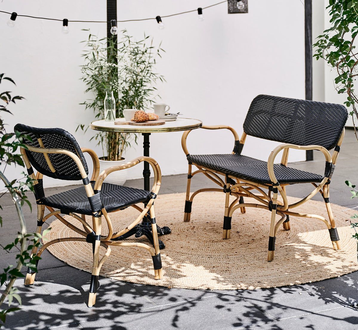 Dette er et livsstilsbillede af Bistro Bench i skin on Rattan frame, i et udendørs café-miljø med et bistrobord på mørke fliser.