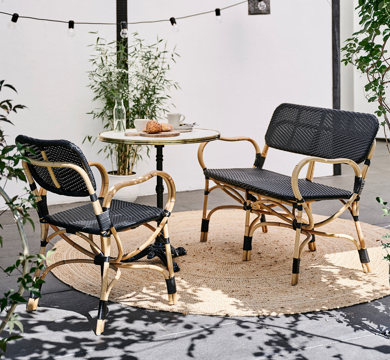 Dette er et livsstilsbillede af Bistro Bench i skin on Rattan frame, i et udendørs café-miljø med et bistrobord på mørke fliser.