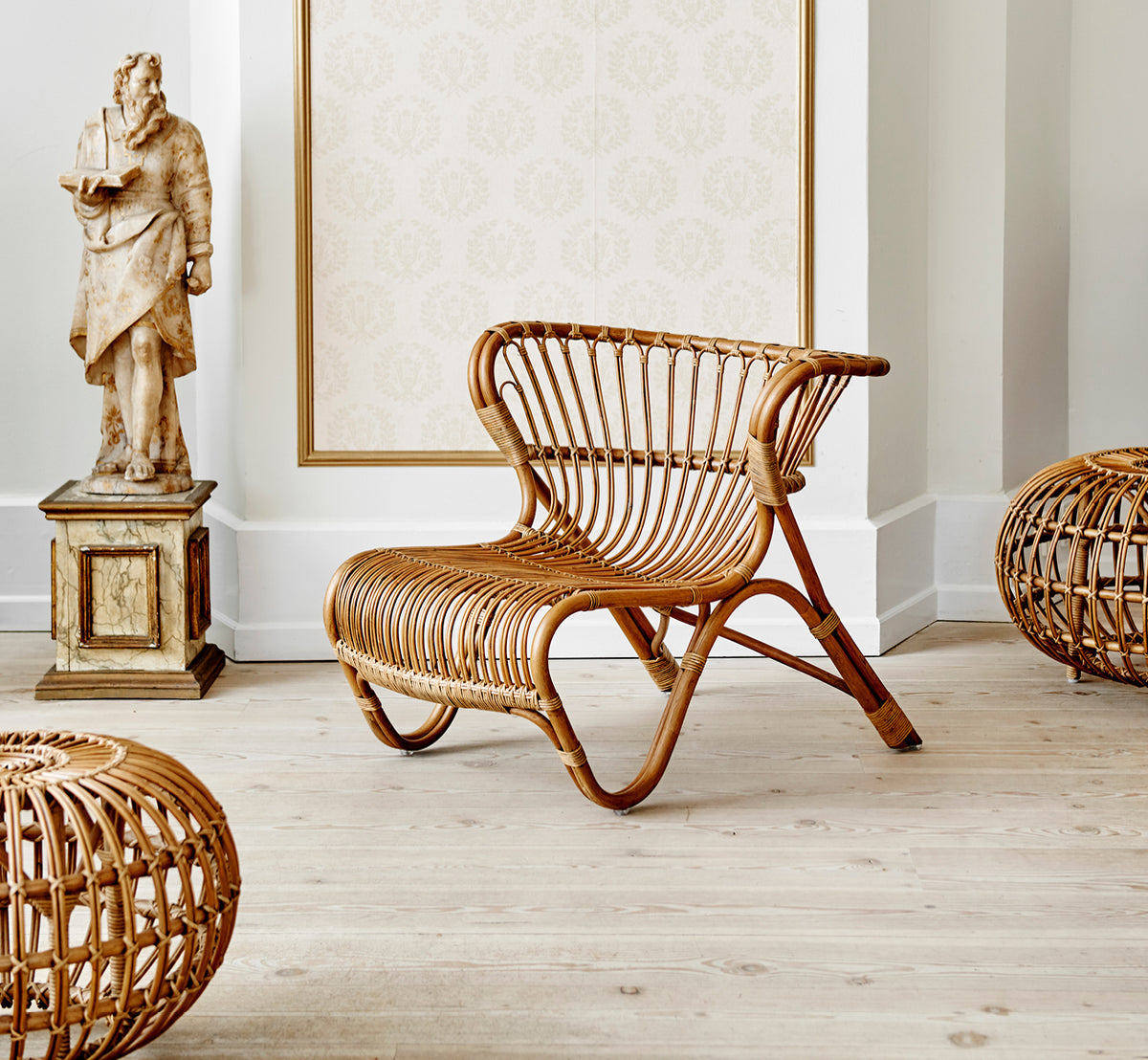 Dette er et livsstilsbillede af Ottoman Small i polished antique rattan. Placeret i et rum med udsmykket tapet og en klassisk statue.