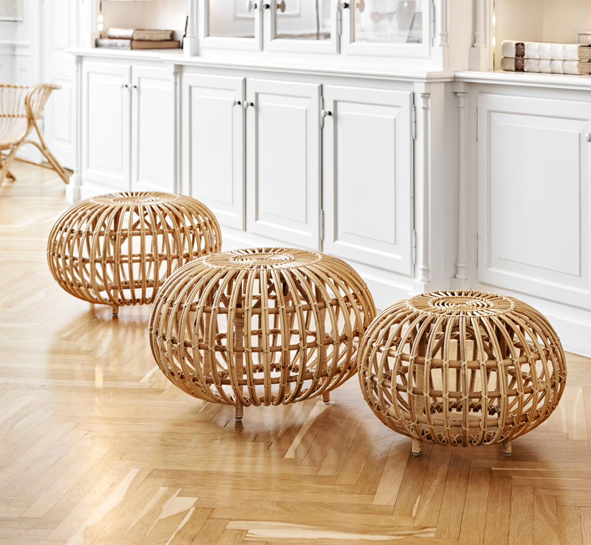 Dette er et livsstilsbillede af Ottoman Small i natural skin on rattan. Samlet i et klassisk panelinteriør med et antikt ur.