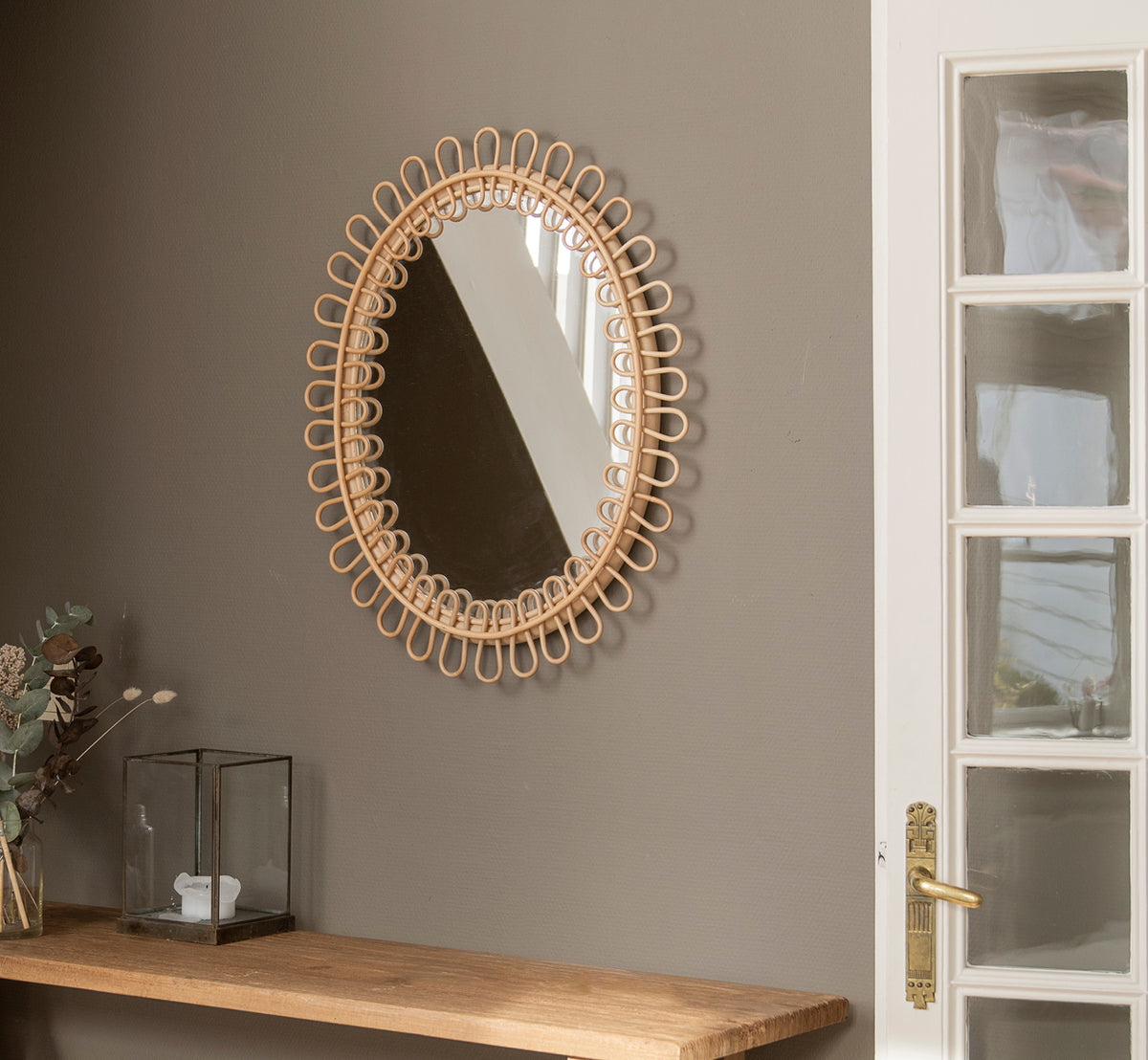 Dette er et livsstilsbillede af Luella Spejl i polished natural rattan. Hængende på en grå væg over en træhylde med eukalyptus og glasdekoration.
