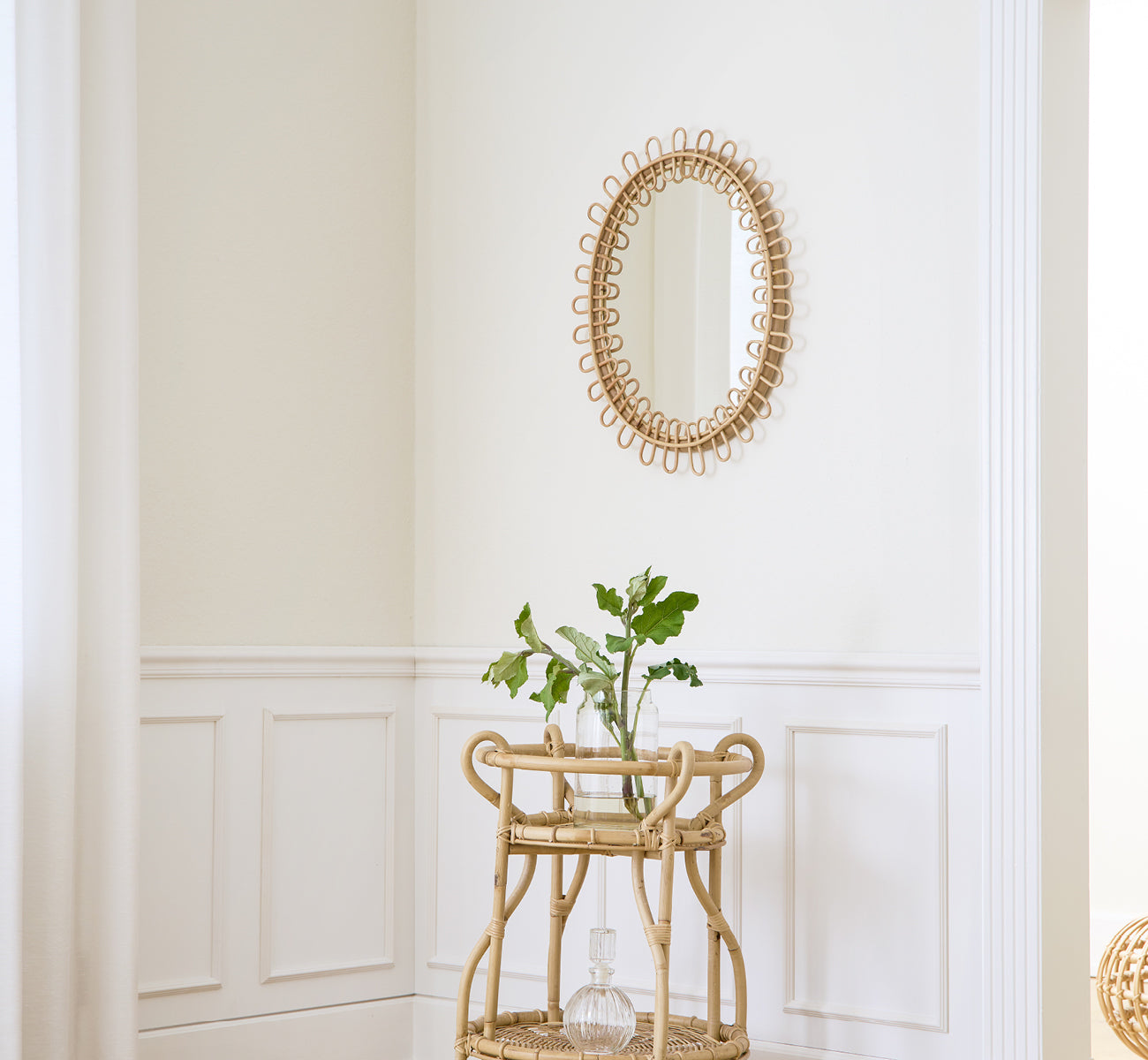 Dette er et livsstilsbillede af Luella Spejl i polished natural rattan. Monteret på en hvid panelvæg over en rattan-barvogn med grønt.
