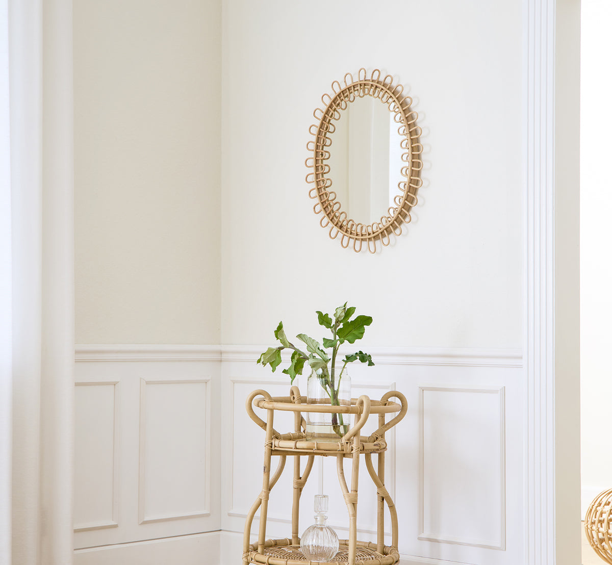 Dette er et livsstilsbillede af Luella Spejl i polished natural rattan. Monteret på en hvid panelvæg over en rattan-barvogn med grønt.