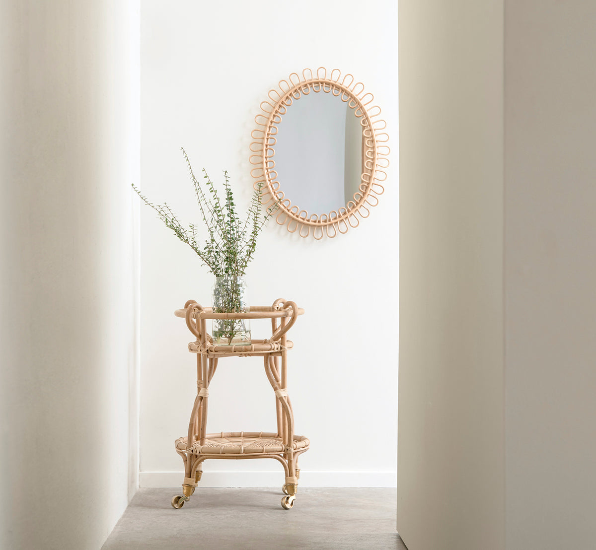 Dette er et livsstilsbillede af Luella Spejl i polished natural rattan. I en lys entré-niche over en rattan-barvogn med friskt grønt.