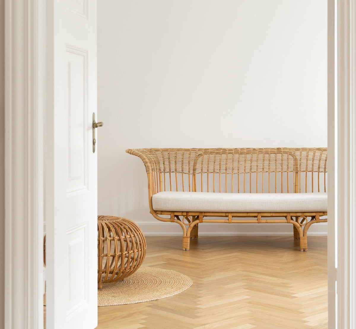 Dette er et livsstilsbillede af Belladonna Sofa i polished natural rattan, i et lyst skandinavisk interiør set gennem en hvid dørramme.