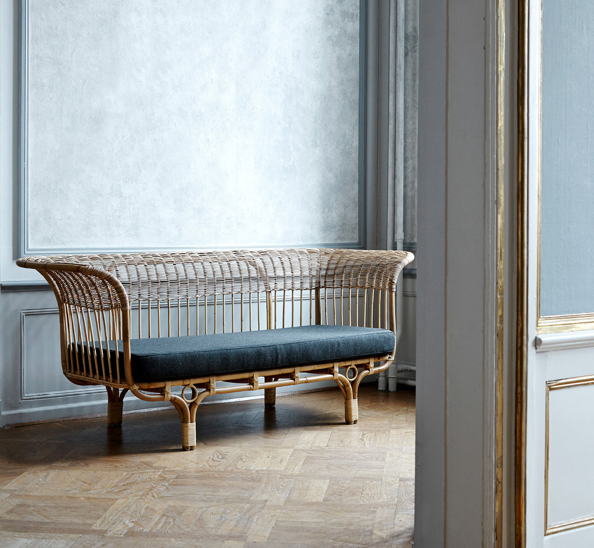 Dette er et livsstilsbillede af Belladonna Sofa i polished natural rattan, i et lyst skandinavisk interiør set gennem en hvid dørramme.