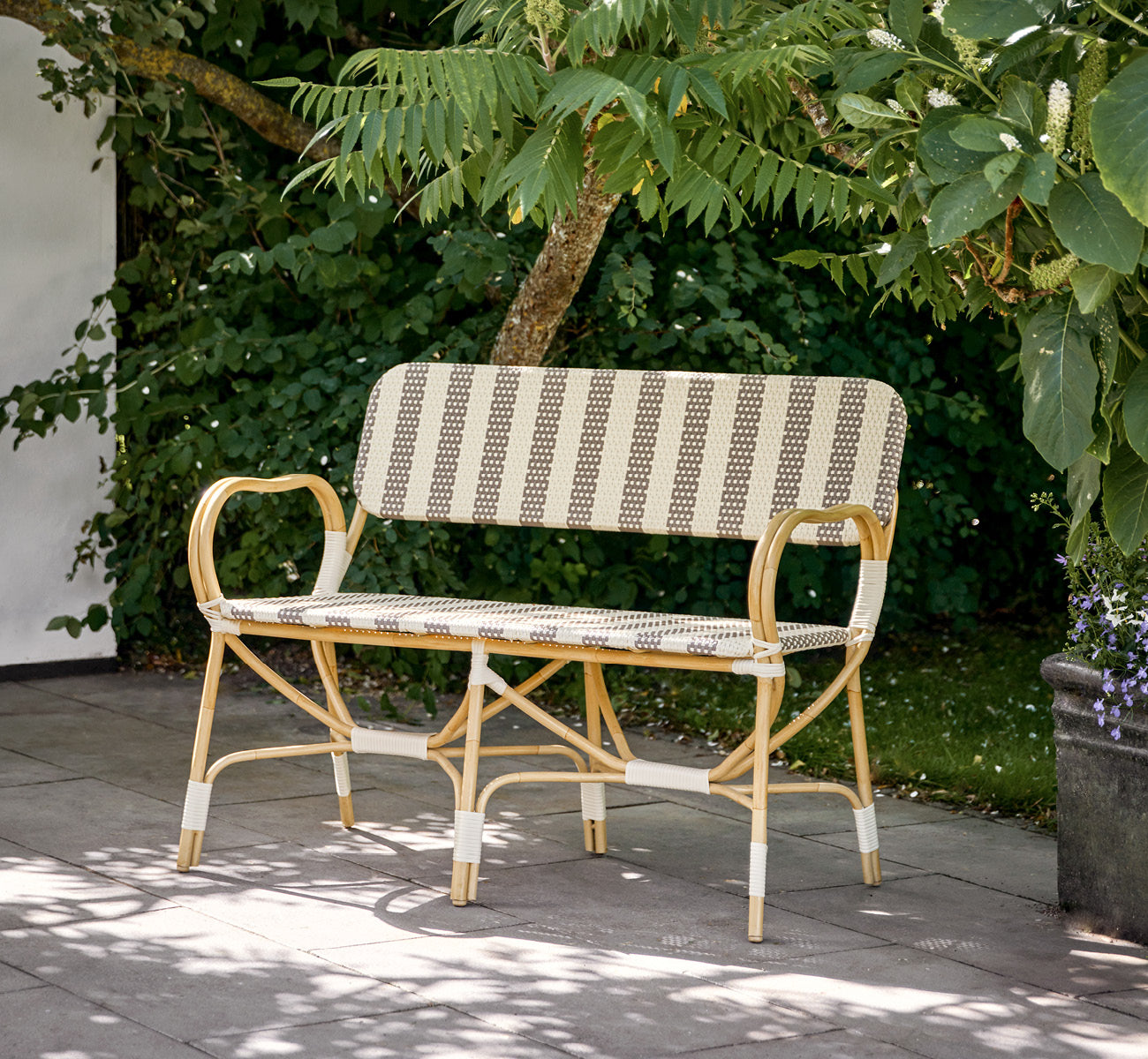 Dette er et livsstilsbillede af Bistro Exterior Bænk i natural Aluminium ArtFibre, i et udendørs havecafé med et bistrobord og omkringstående træer.