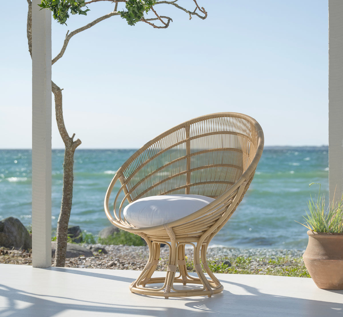 Dette er et livsstilsbillede af Luna Udendørs lænestol i natural aluminium artfibre. På en hvid strandterrasse med havudsigt og et træ.
