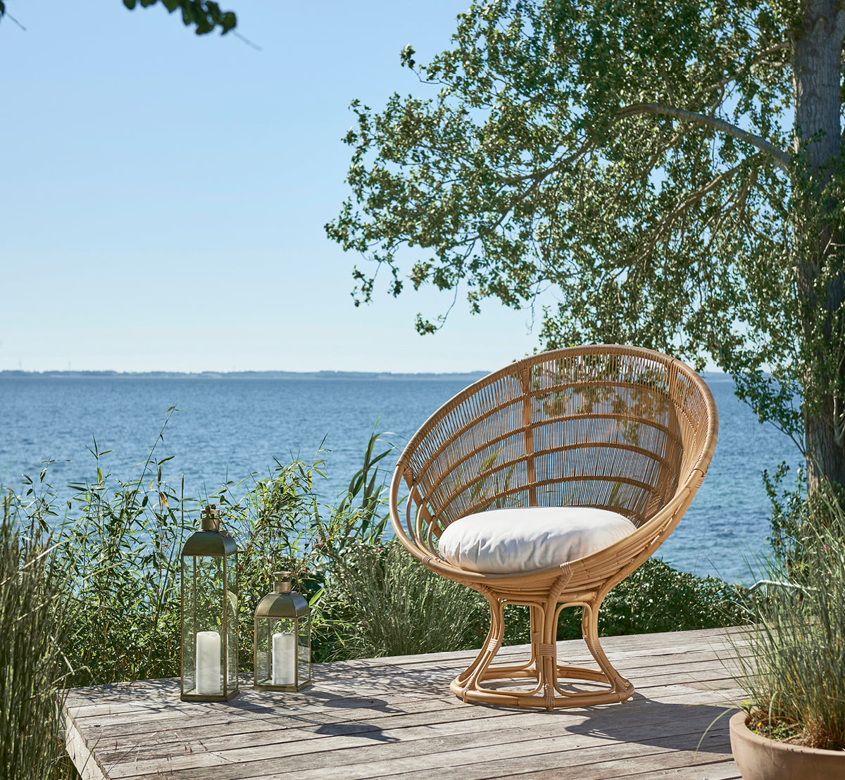 Dette er et livsstilsbillede af Luna Udendørs lænestol i natural aluminium artfibre. På en trædæk ved havet omgivet af frodig beplantning.