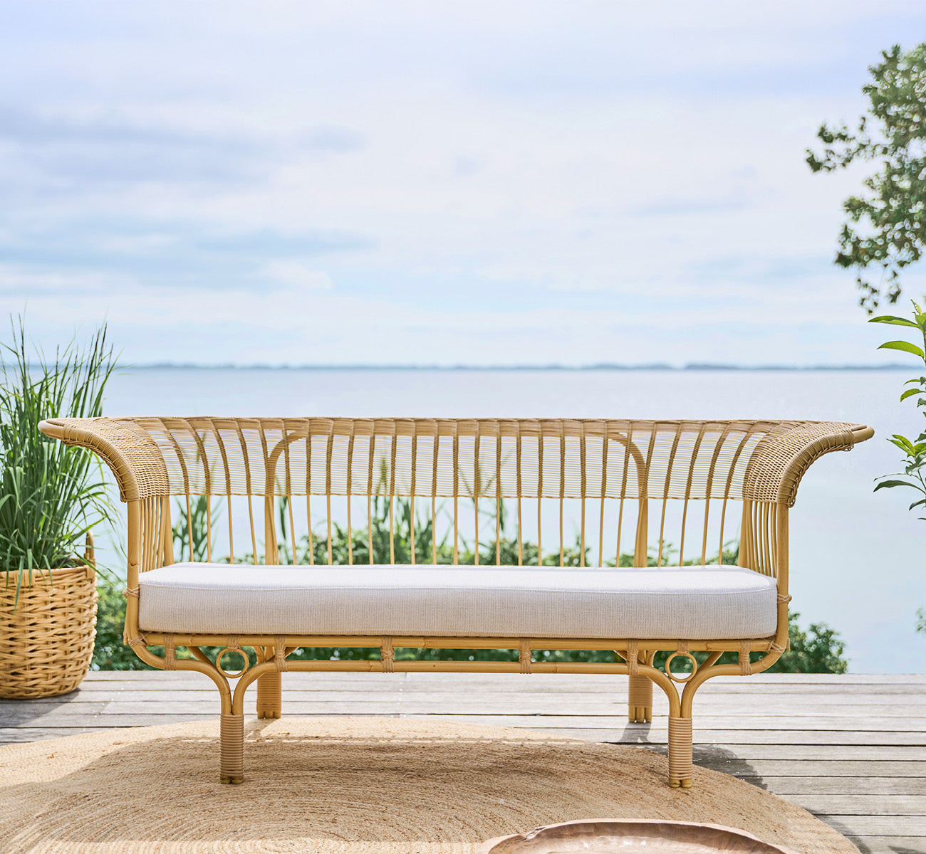Dette er et livsstilsbillede af Belladonna Udendørs sofa i natural Aluminium ArtFibre, på et udendørs trædæk med udsigt over vandet.