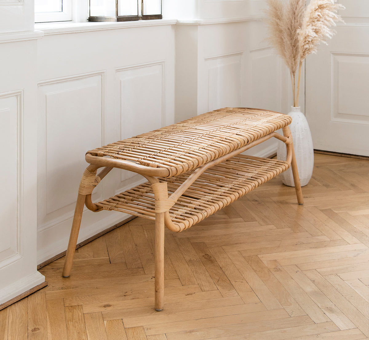 Dette er et livsstilsbillede af Machiya Bænk i natural skin on rattan. Hvid panelgang med sildebensgulv og vase med pampasgræs.