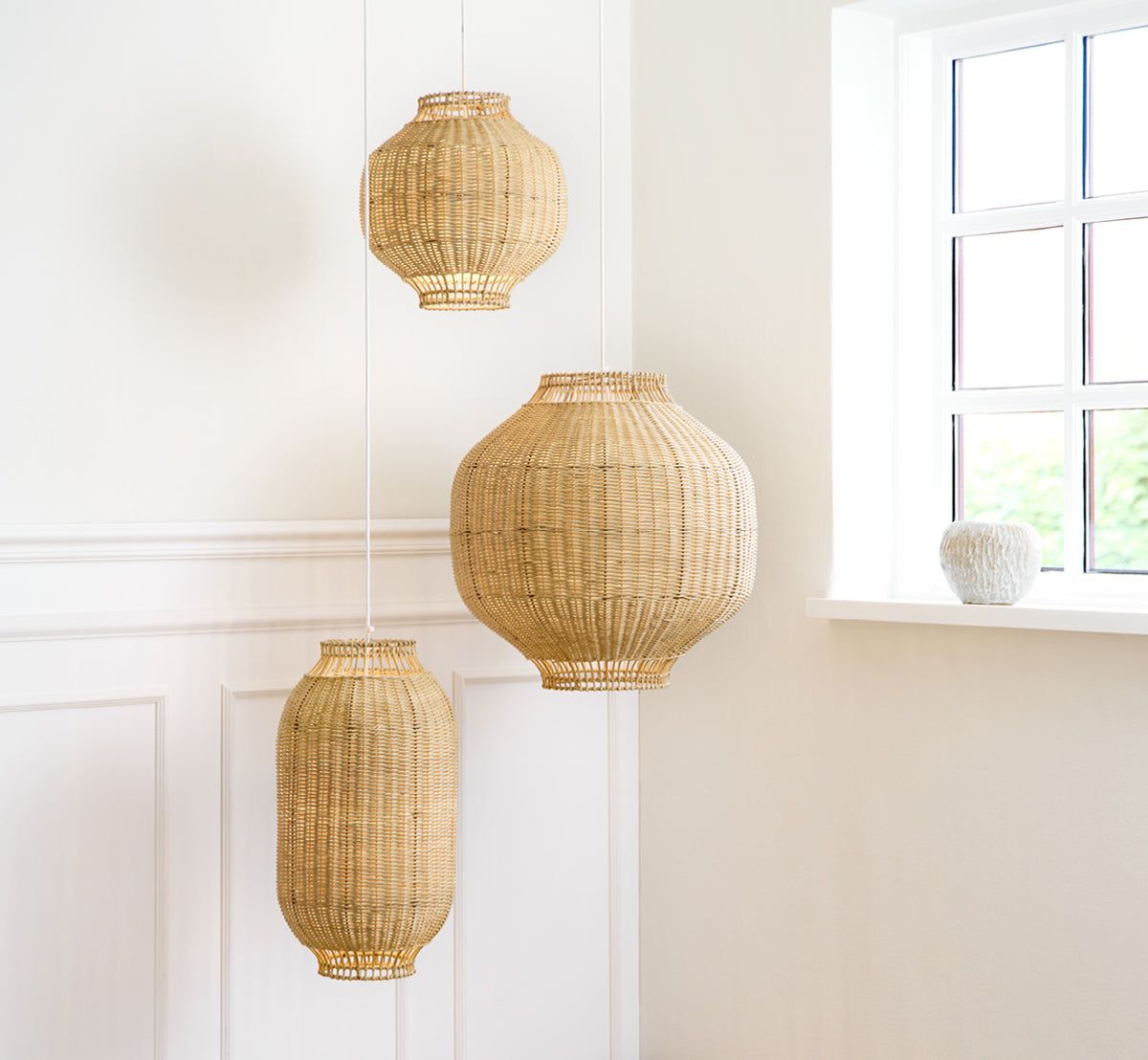 Dette er et livsstilsbillede af Kou Lampeskærm i natural core weave rattan frame. Tre størrelser hænger sammen i et lyst hvidt panelrum.