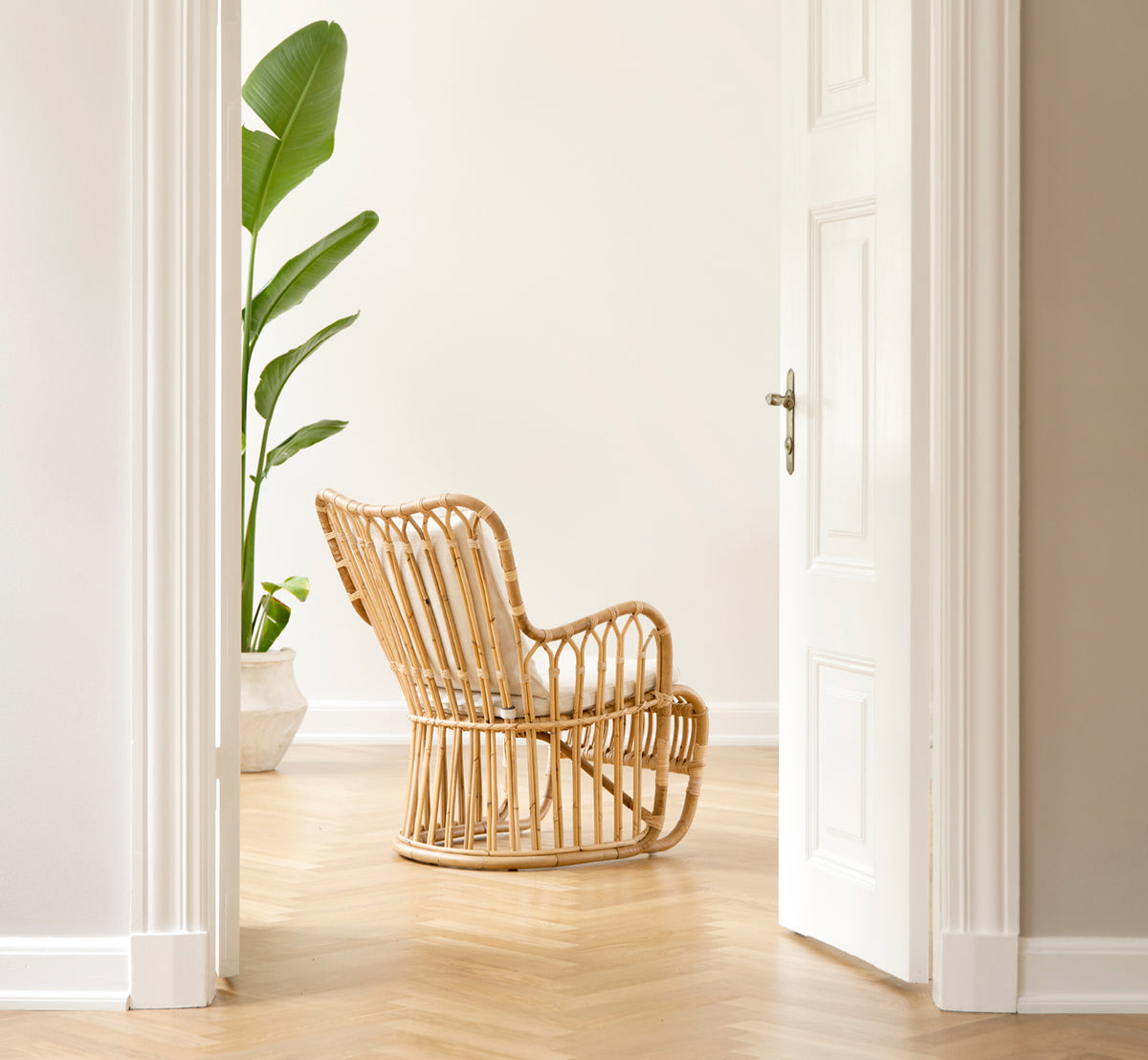 Dette er et livsstilsbillede af Tulip Lænestol i polished natural rattan. Kombineret med en matchende sofa i et lyst panelrum med dør.