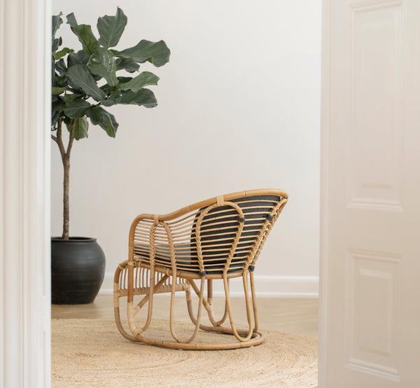 Dette er et livsstilsbillede af Swing Lænestol i polished natural rattan. Stående alene ved siden af en løvrig plante på et sildebensparketgulv.