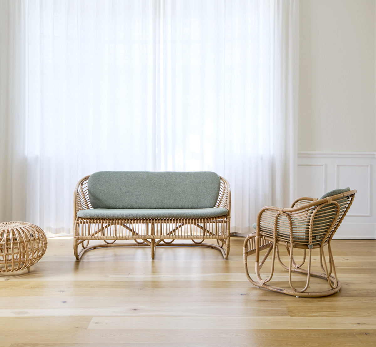 Dette er et livsstilsbillede af Swing Sofa i polished natural rattan. Placeret på et tæppe ved siden af en fiddle-leaf plante på parketgulv.