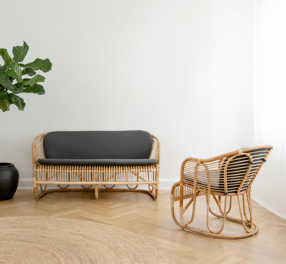 Dette er et livsstilsbillede af Swing Sofa i polished natural rattan. Placeret på et tæppe ved siden af en fiddle-leaf plante på parketgulv.