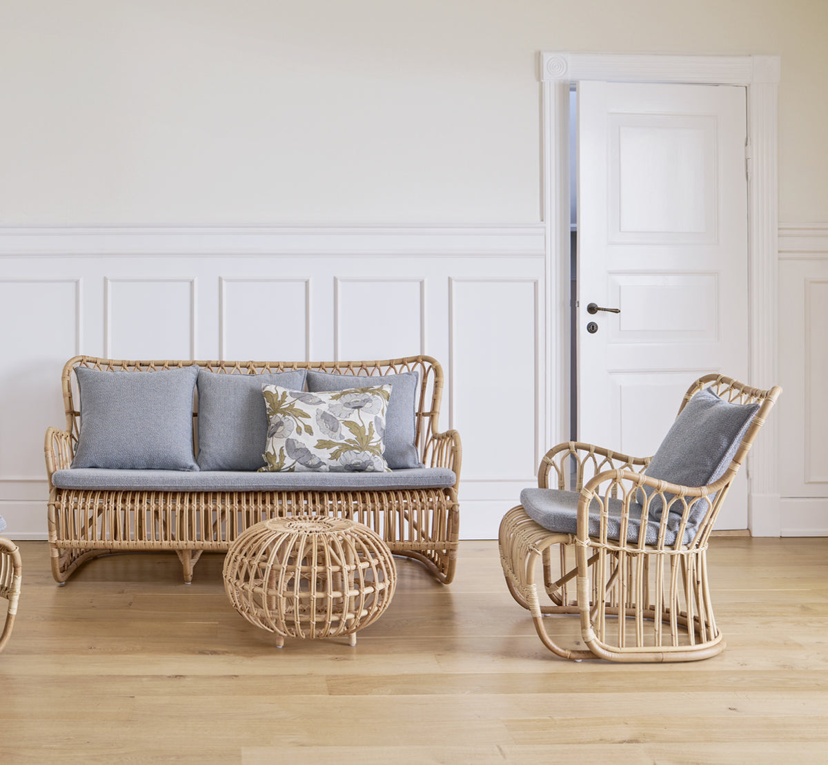 Dette er et livsstilsbillede af Tulip Sofa i polished natural rattan. Kombineret med en rattan-lænestol ved et solbeskinnet gardinvindue.