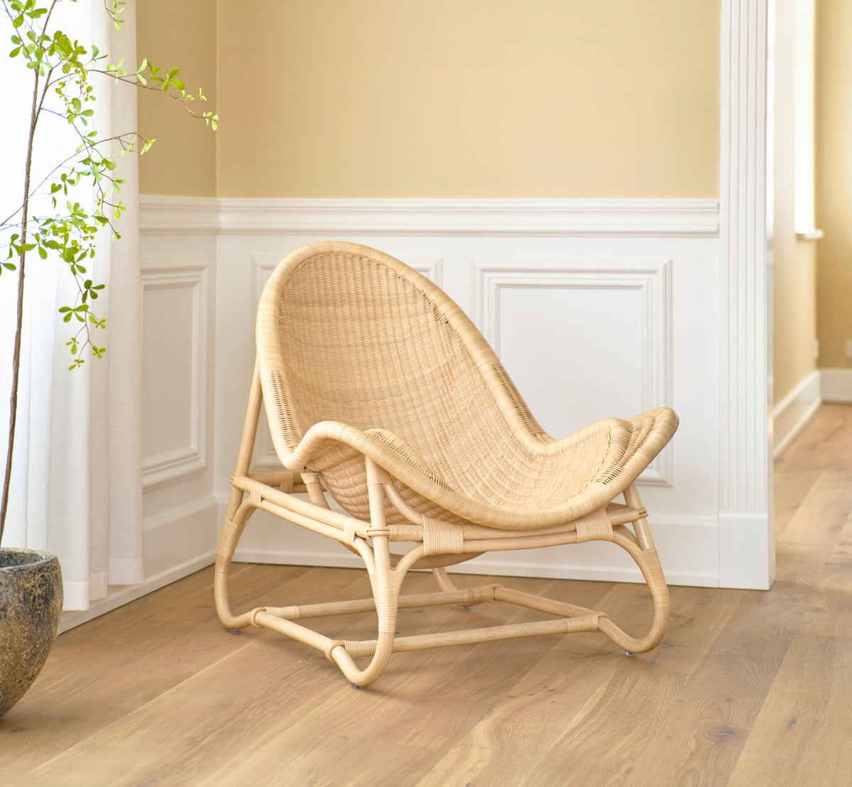 Dette er et livsstilsbillede af Pacifique Lænestol i natural core weave rattan frame. Ved hvide paneler og en potteplante.
