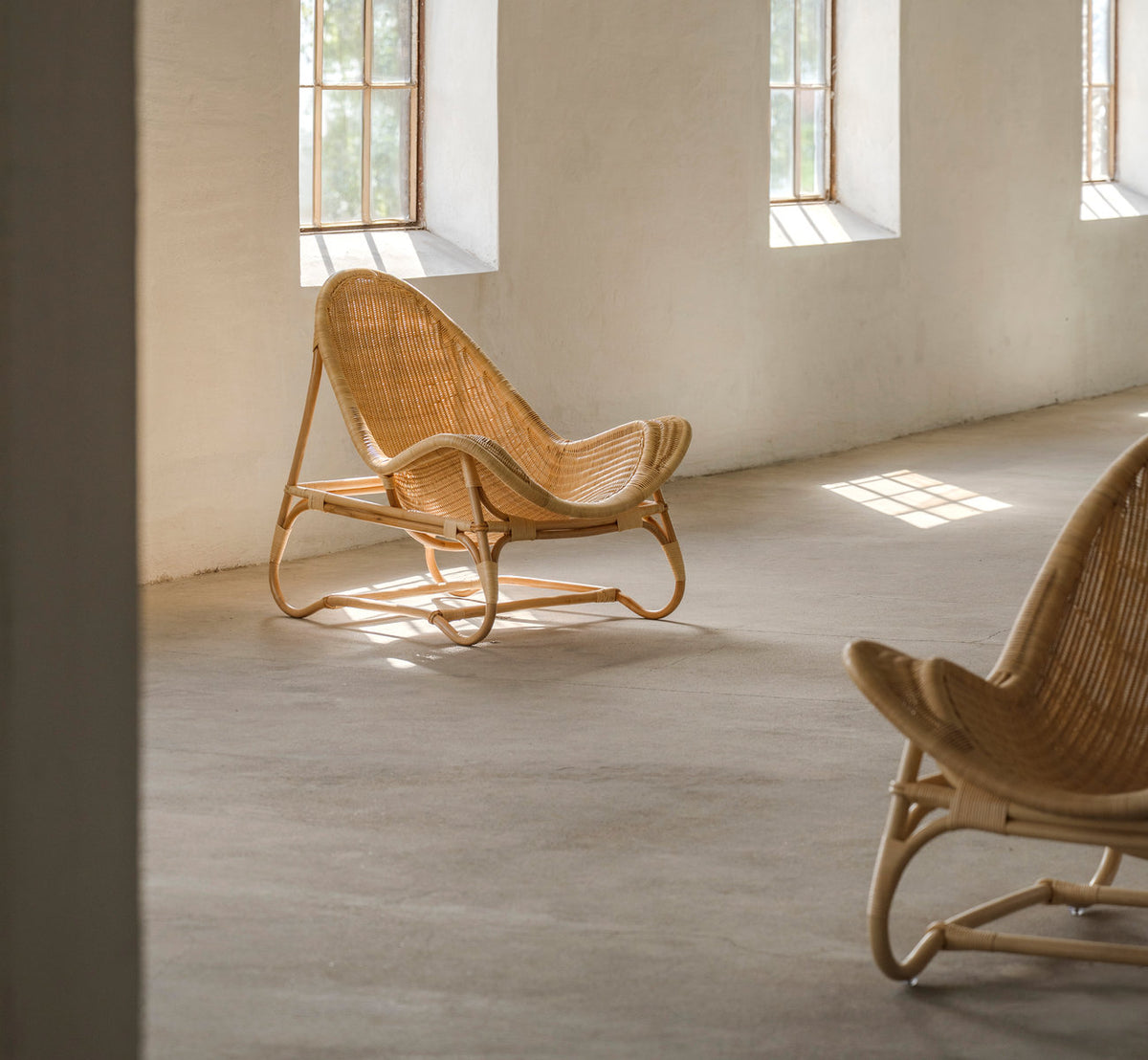 Dette er et livsstilsbillede af Pacifique Lænestol i natural core weave rattan frame. To i et solbeskinnet rum med betongulv.