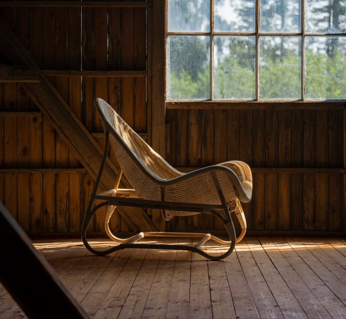 Dette er et livsstilsbillede af Pacifique Lænestol i natural core weave rattan frame. I en træklædt lade med varmt lys fra vinduet.