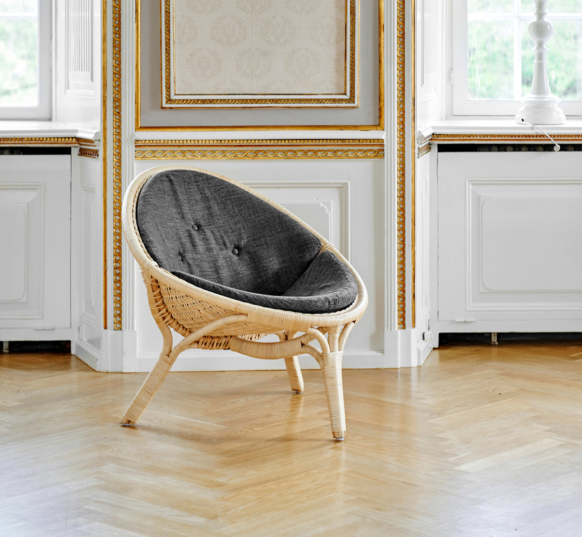 Dette er et livsstilsbillede af Rana Lænestol i natural core weave rattan frame. Stående alene i et rum med forgyldt panel og parketgulv.