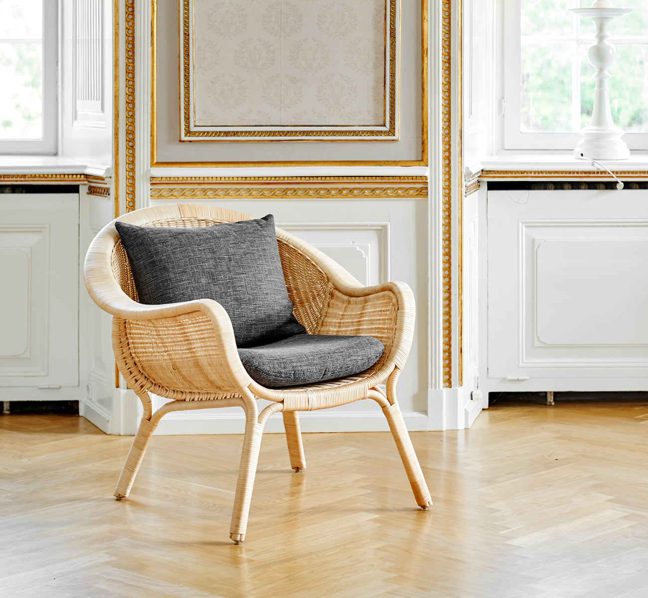 Dette er et livsstilsbillede af Madame Lænestol i natural core weave rattan frame. Elegant forgyldt rum med egegulv og grå pude.