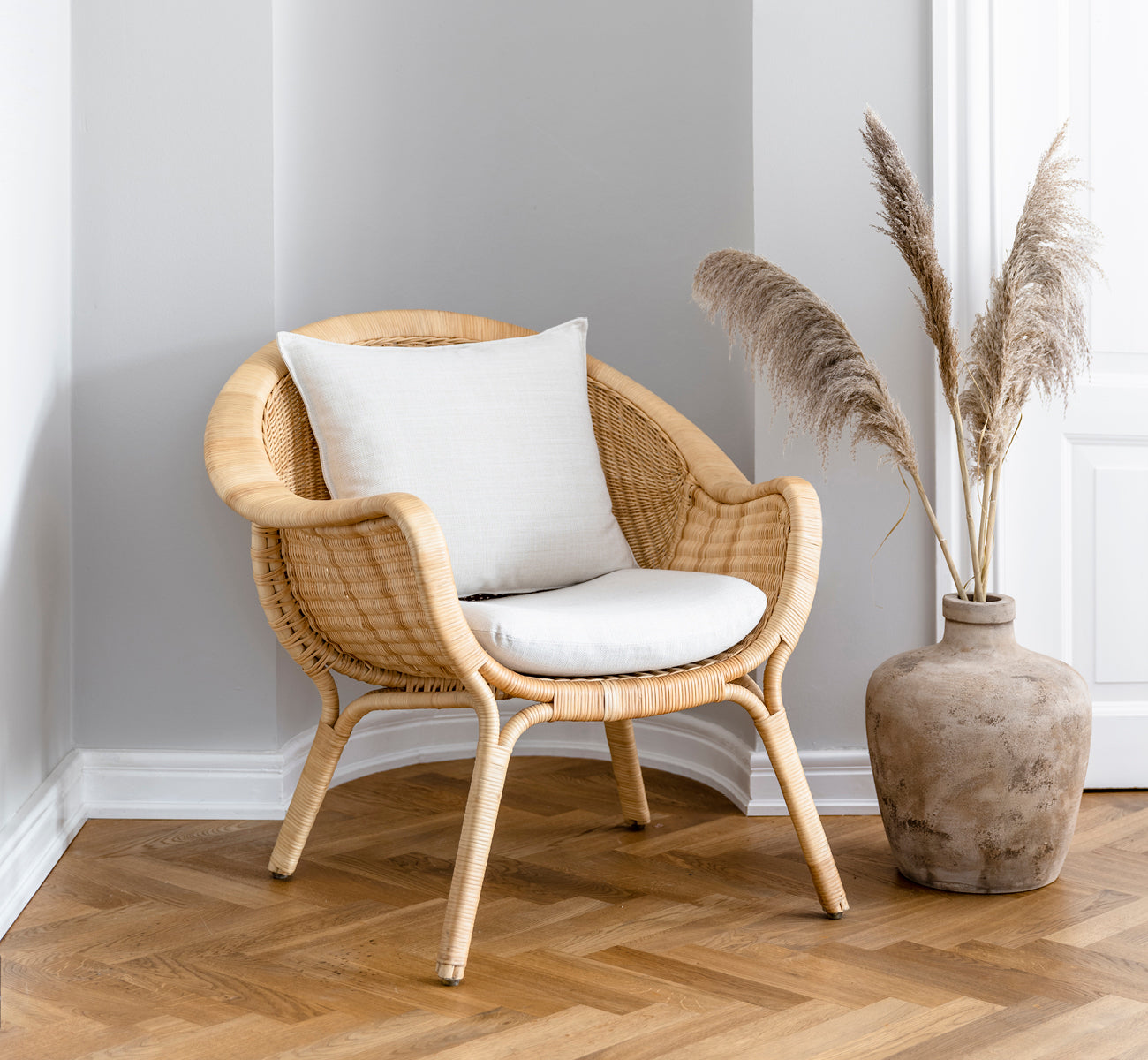 Dette er et livsstilsbillede af Madame Lænestol i natural core weave rattan frame. Hyggeligt hjørne med sildebensgulv og vase med pampasgræs.