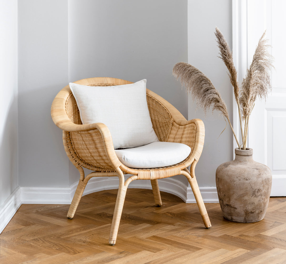 Dette er et livsstilsbillede af Madame Lænestol i natural core weave rattan frame. Hyggeligt hjørne med sildebensgulv og vase med pampasgræs.