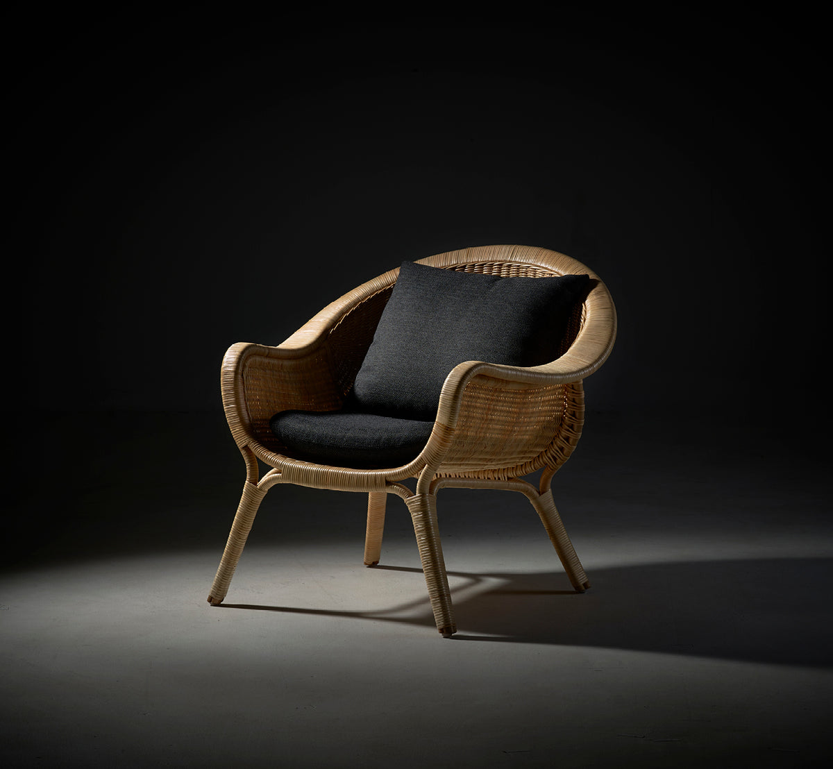 Dette er et livsstilsbillede af Madame Lænestol i natural core weave rattan frame. Dramatisk studielys med mørk pude på grå baggrund.