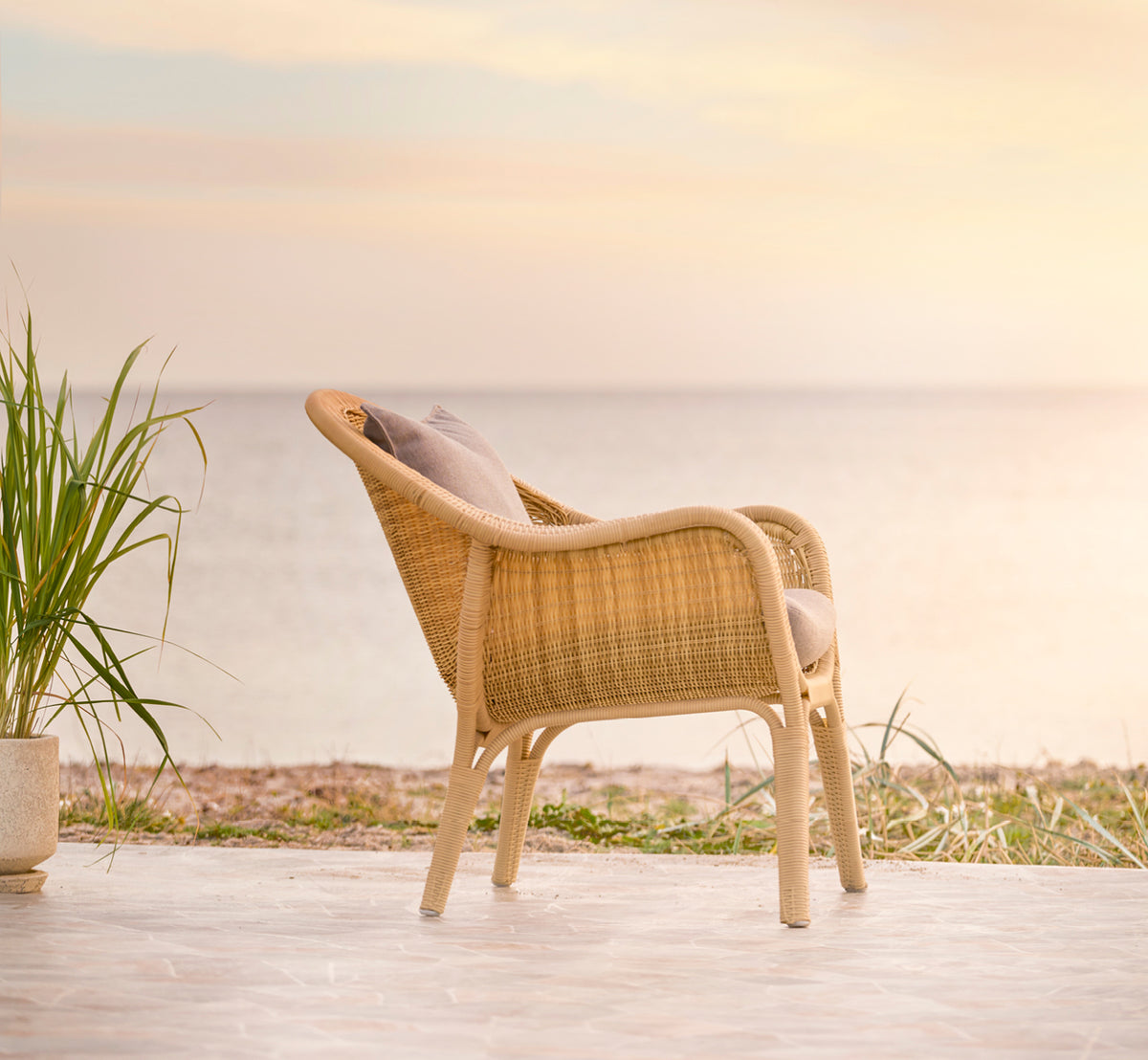 Dette er et livsstilsbillede af Madame Lænestol i natural core weave ArtFibre Aluminium, på en åben kystterrasse ved solnedgang med havudsigt og.