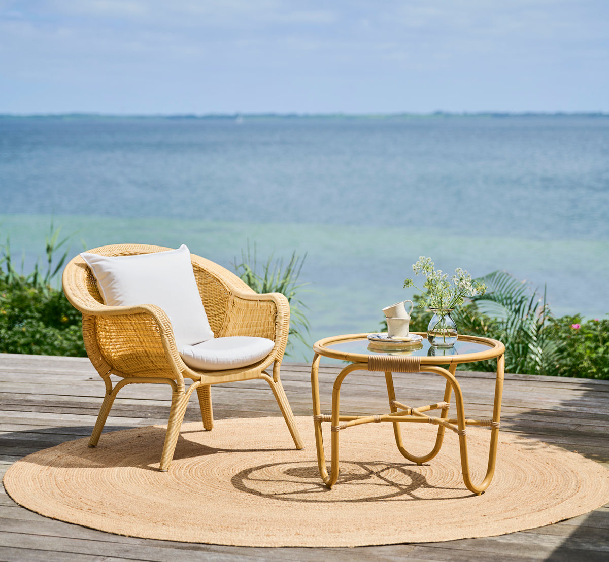 Dette er et livsstilsbillede af Madame Lænestol i natural core weave ArtFibre Aluminium, på en åben kystterrasse ved solnedgang med havudsigt og.
