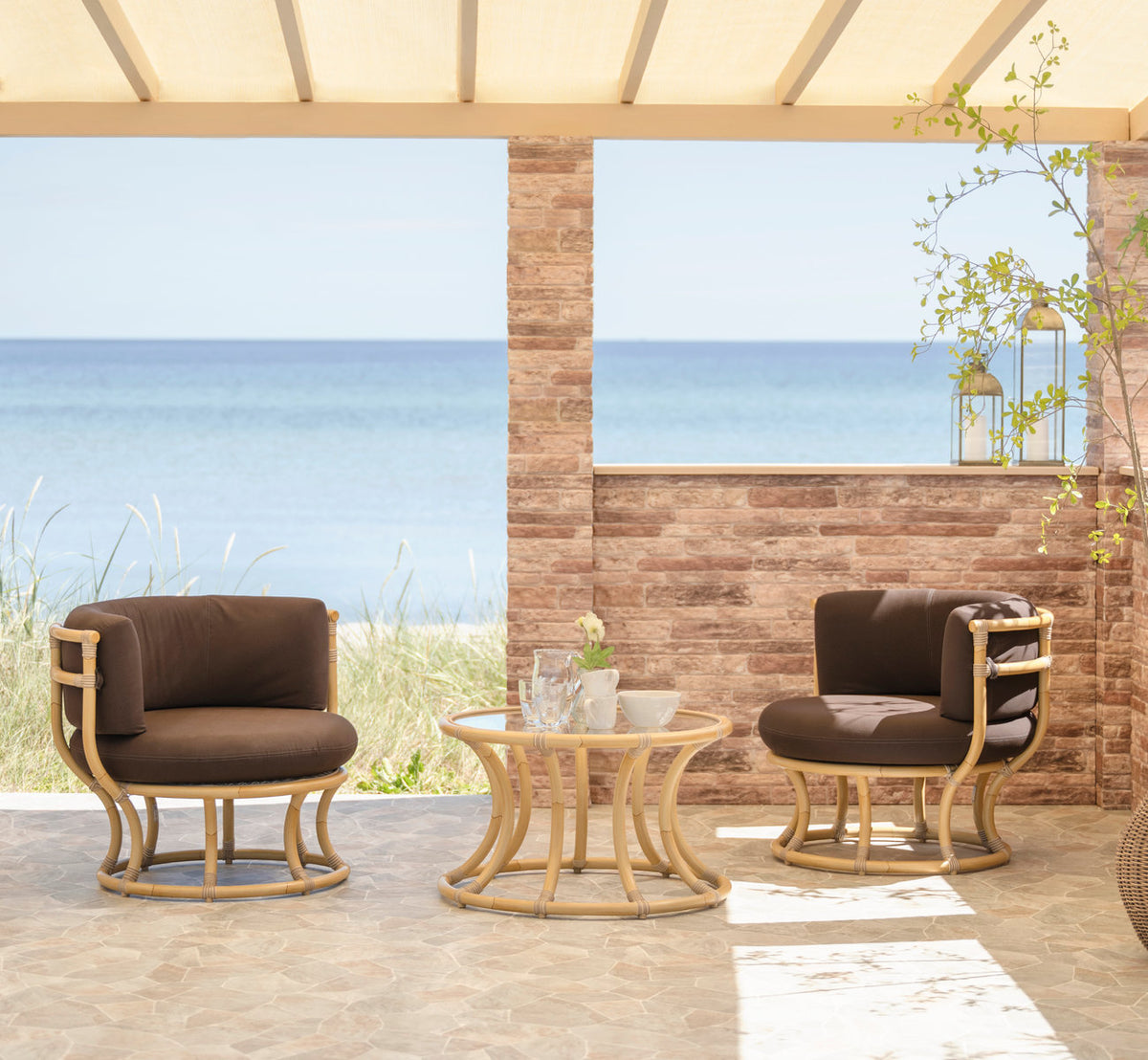 Dette er et livsstilsbillede af Coco Udendørs lænestol i natural aluminium artfibre. Lænestole og bord på en terrasse ved havet.