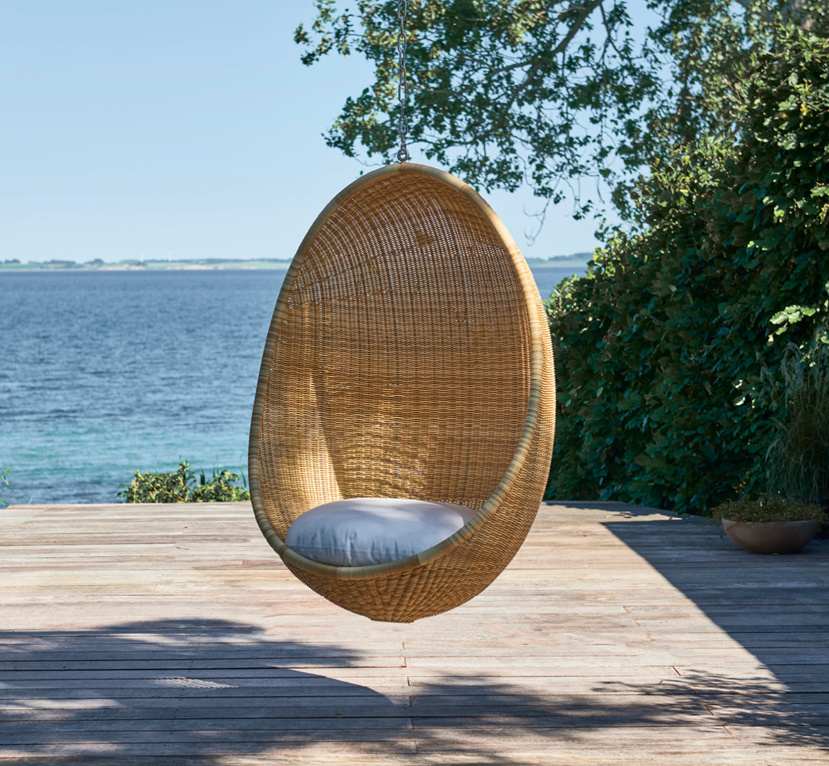 Dette er et livsstilsbillede af Hanging Udendørs hængeæg-stol i natural core weave ArtFibre Aluminium, hængende fra en hvid pergola med sandstrand og.