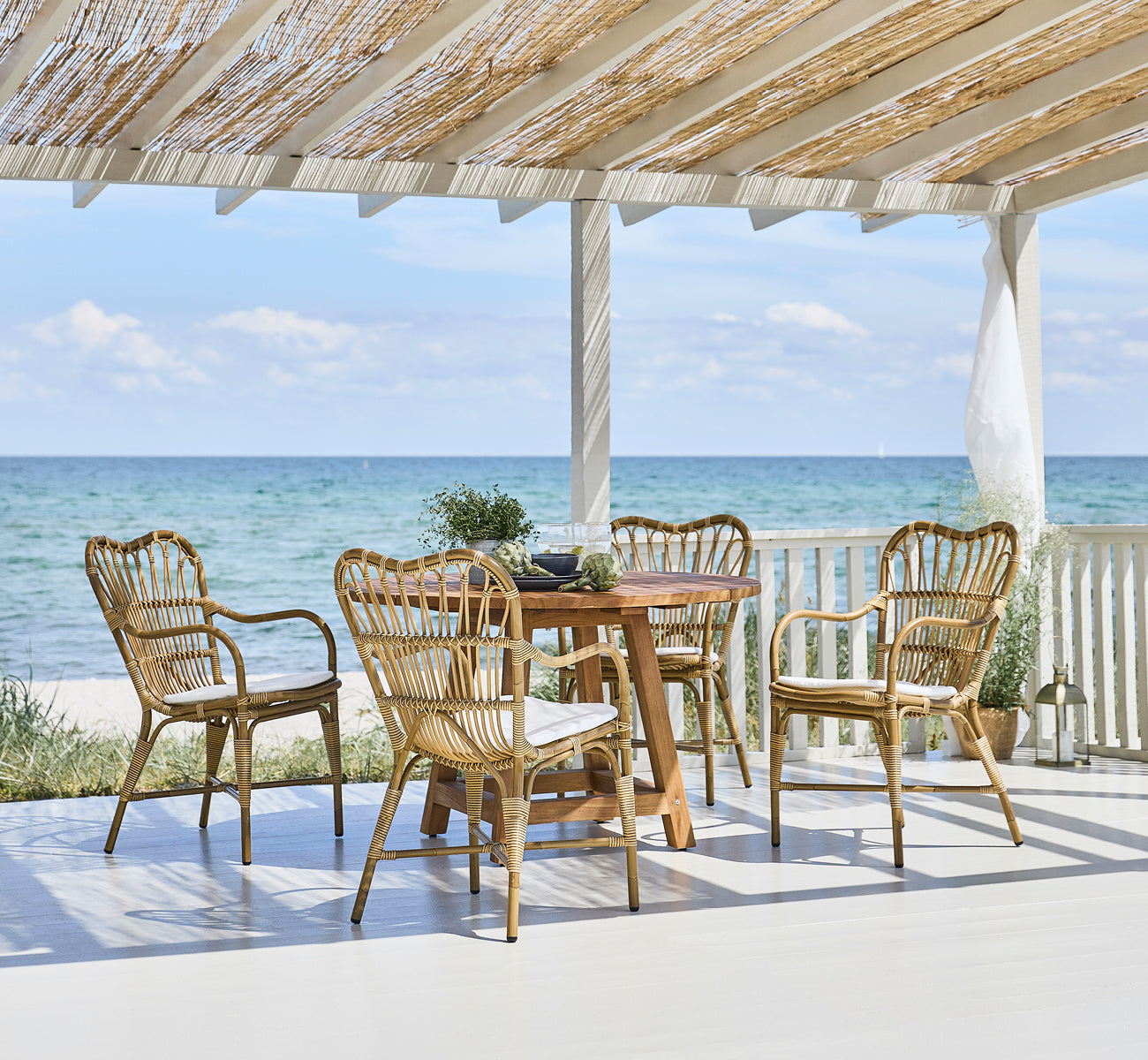 Dette er et livsstilsbillede af Margret Udendørs spisestuelænestol i antique aluminium artfibre. Strandpavillon med teakbord og havudsigt.