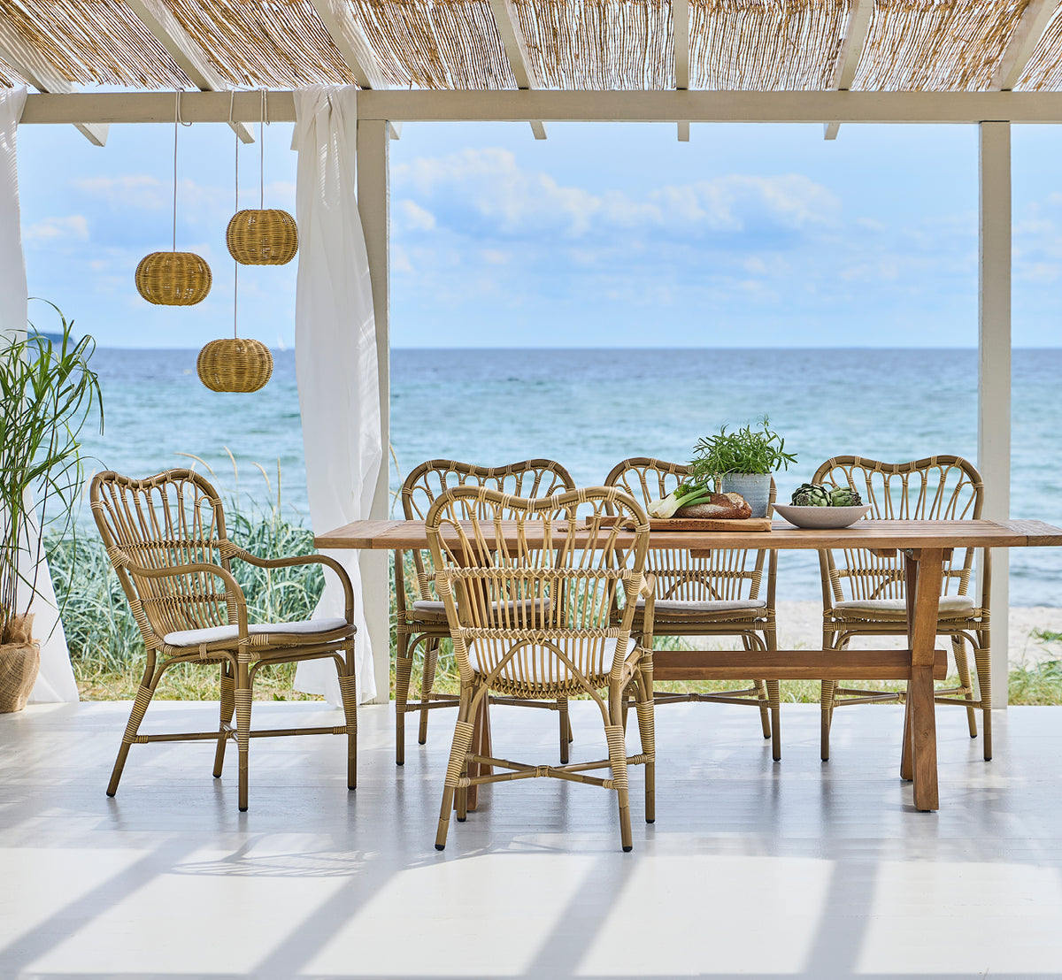 Dette er et livsstilsbillede af Margret Udendørs spisestuelænestol i antique aluminium artfibre. Strandpavillon med pendellamper og havudsigt.