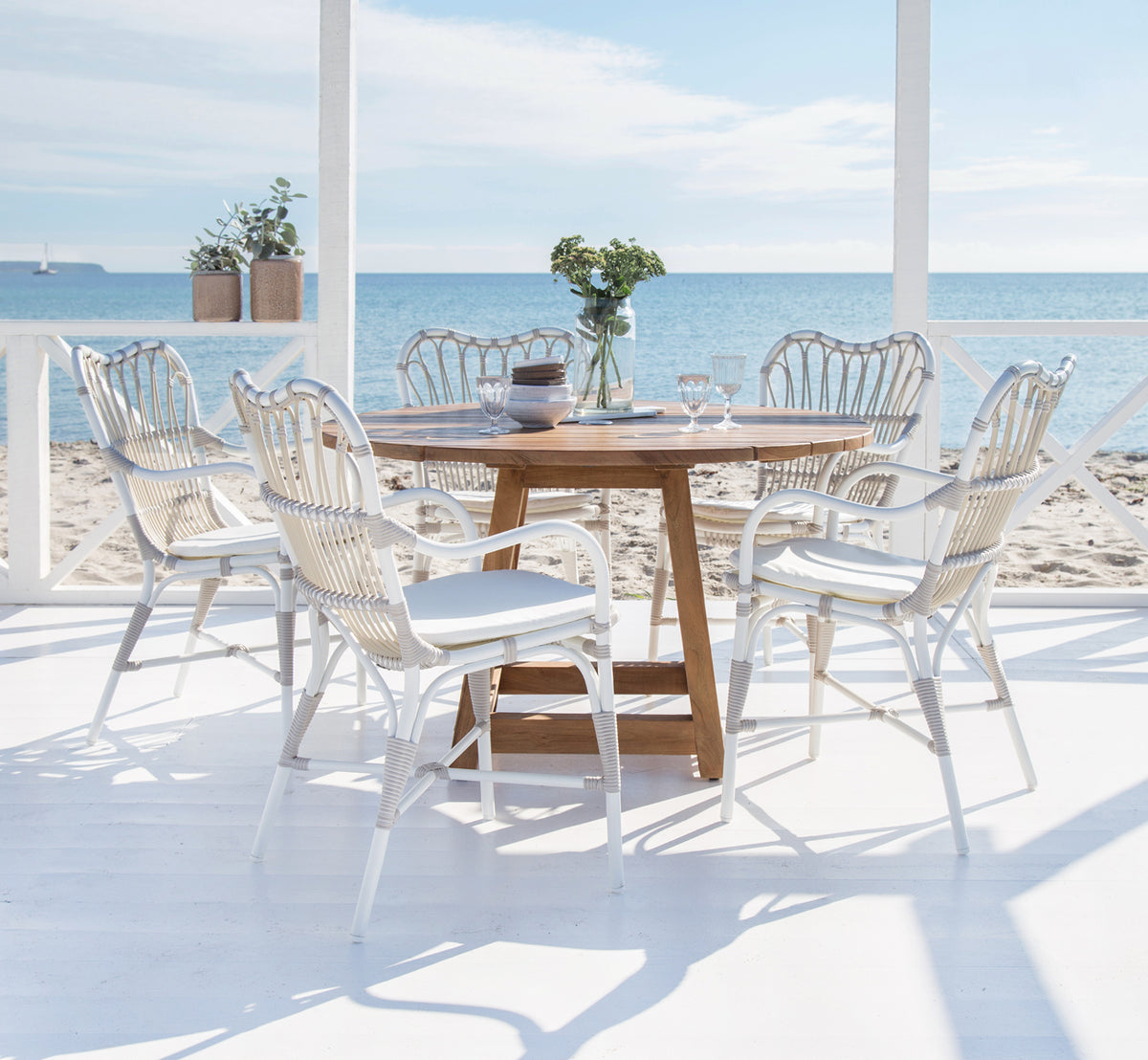Dette er et livsstilsbillede af Margret Udendørs spisestuelænestol i dove white aluminium artfibre. Strandterrasse med teakbord og havudsigt.