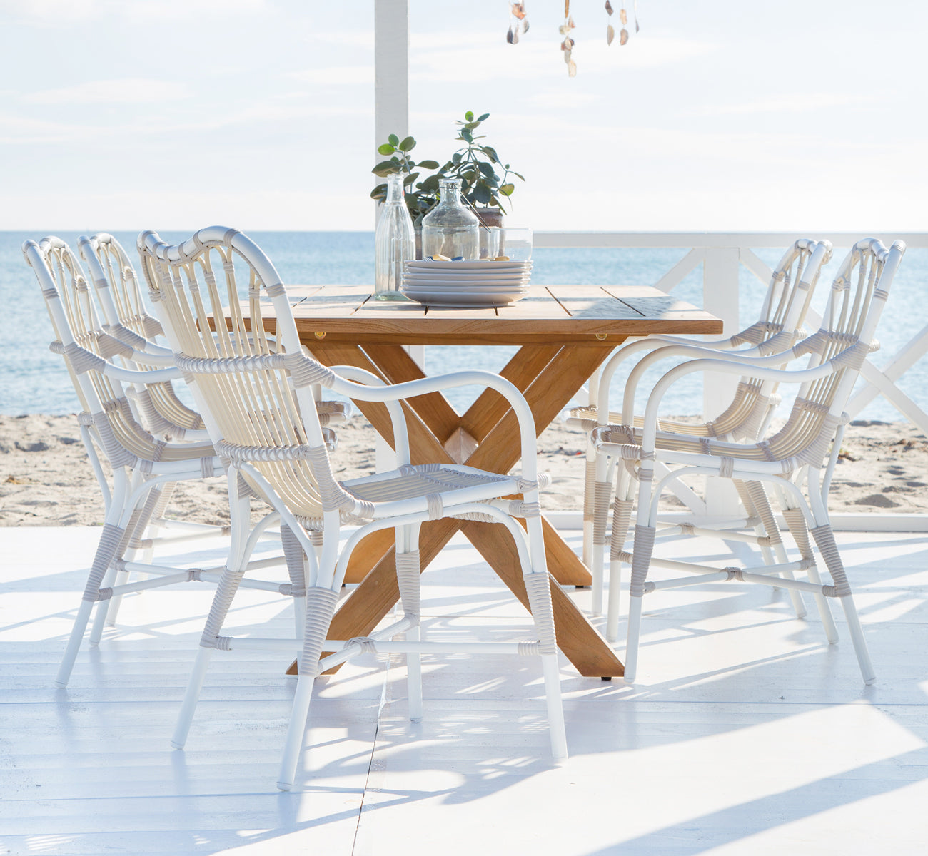 Dette er et livsstilsbillede af Margret Udendørs spisestuelænestol i dove white aluminium artfibre. Strandterrasse med teakbord og dekorationer.