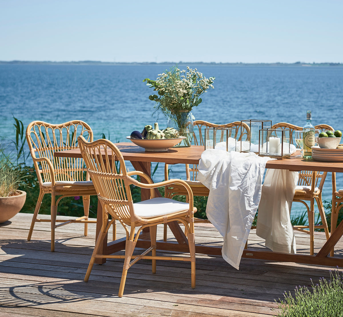 Dette er et livsstilsbillede af Margret Udendørs spisestuelænestol i natural aluminium artfibre. Trædæk ved havet med teakbord og blomster.