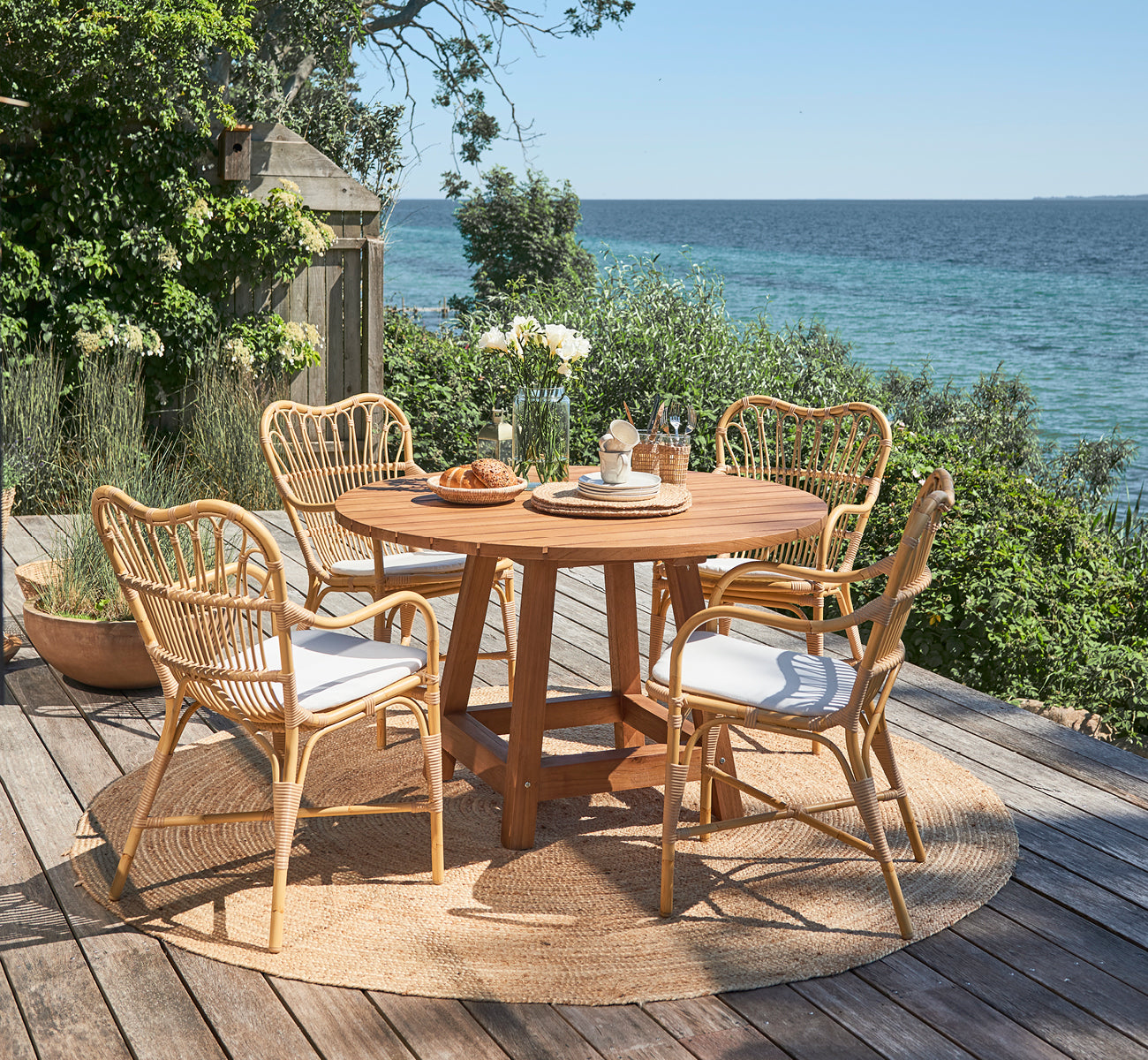 Dette er et livsstilsbillede af Margret Udendørs spisestuelænestol i natural aluminium artfibre. Dæk ved havet med rundt teakbord og haveplanter.