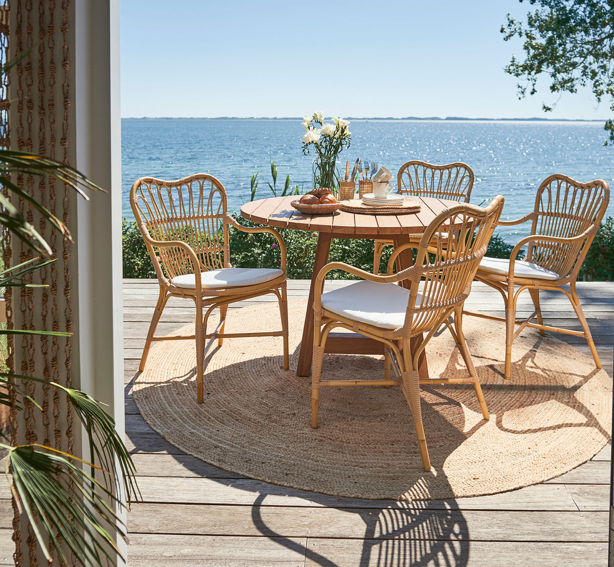 Dette er et livsstilsbillede af Margret Udendørs spisestuelænestol i natural aluminium artfibre. Dæk ved havet med rundt teakbord og havudsigt.